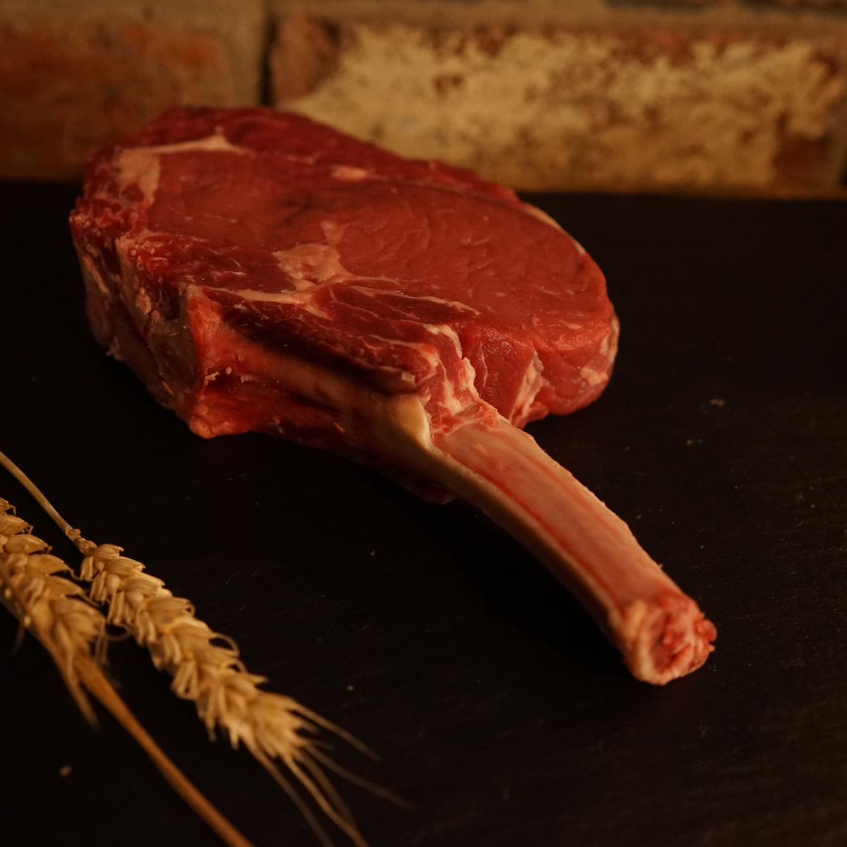 A fresh Aberdeen Angus Tomahawk steak displayed on a dark surface, accompanied by wheat ears, emphasizing its quality and freshness.