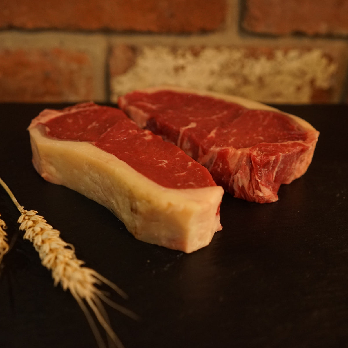 Two fresh Aberdeen Angus sirloin steaks on a dark surface, with a bundle of wheat in the background.