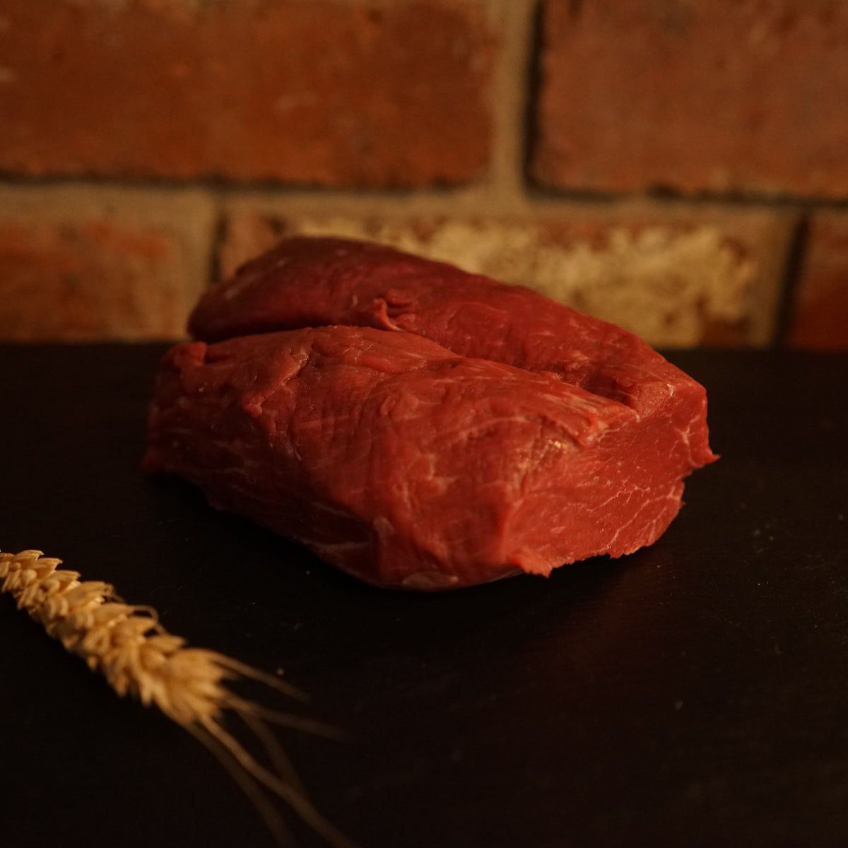 A fresh piece of Aberdeen Angus Chateaubriand steak placed on a dark surface with a brick wall in the background and a wheat ear to the side.