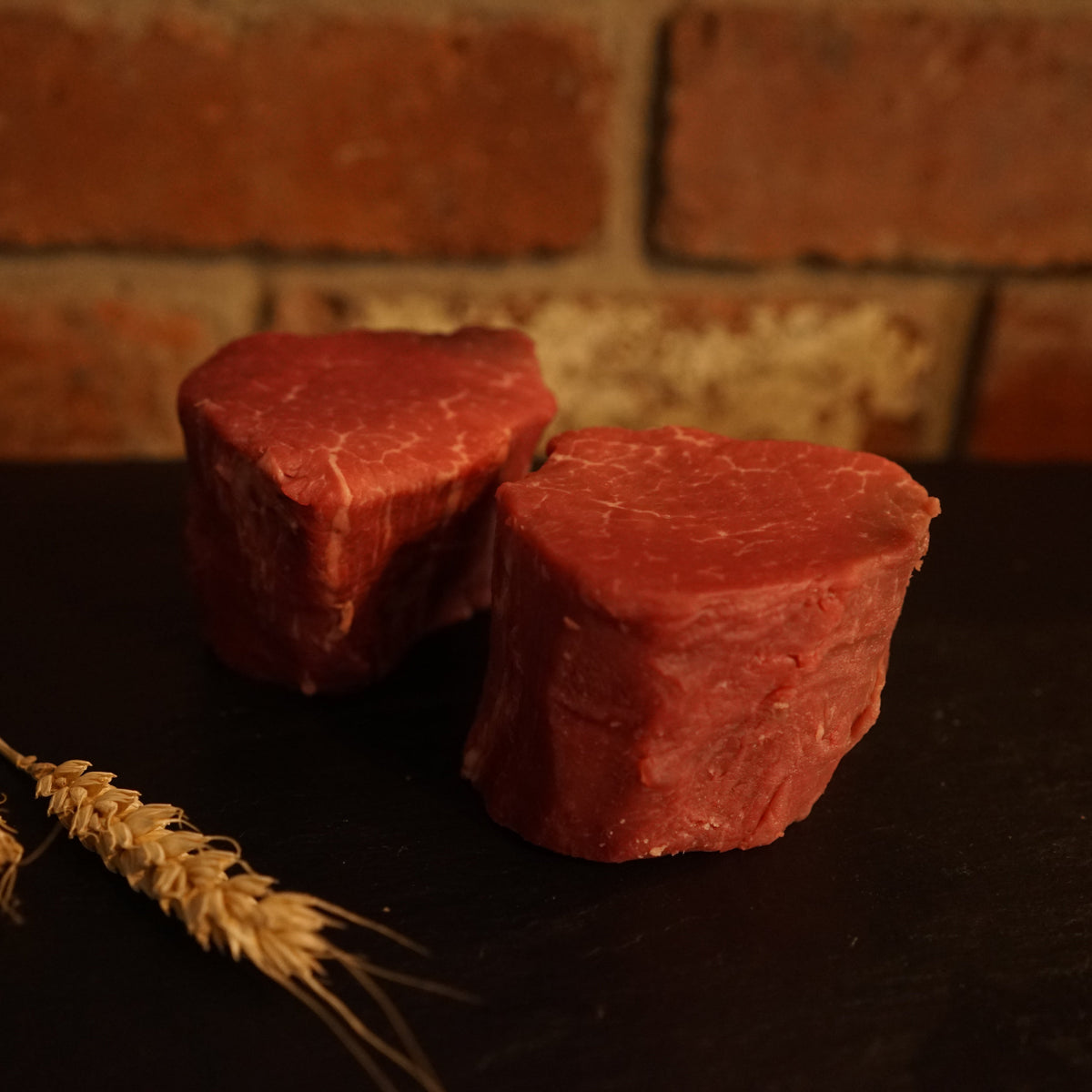 Two fresh Aberdeen Angus fillet steaks on a dark surface with a brick wall in the background, accompanied by two stalks of wheat.