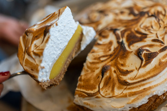 A close-up of a slice of lemon meringue pie being held on a spoon, showing a golden lemon filling, white meringue topping with toasted brown swirls, and a graham cracker crust. A hand with red nail polish is visible holding the spoon.